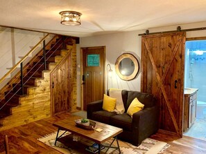 Una habitación con una escalera de madera, un sofá con cojines amarillos, una mesa de centro, un espejo y un baño visible a través de una puerta.