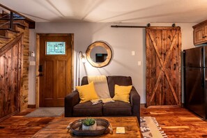Una sala de estar acogedora con puerta de madera, espejo redondo, sofá con cojines y mesa de centro con planta.