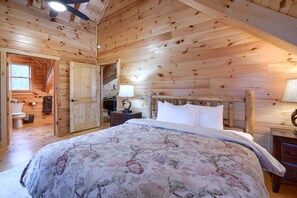 Un dormitorio de cabaña de madera con una cama, mesitas de noche, una lámpara y una puerta que da a un baño.