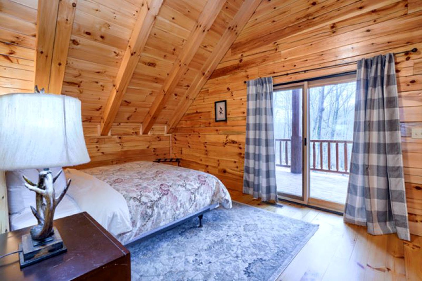 Un dormitorio de cabaña de madera con una cama, una mesita de noche, una lámpara y vistas al exterior a través de un ventanal.