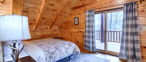 Un dormitorio de cabaña de madera con una cama, una mesita de noche, una lámpara y vistas al exterior a través de un ventanal.
