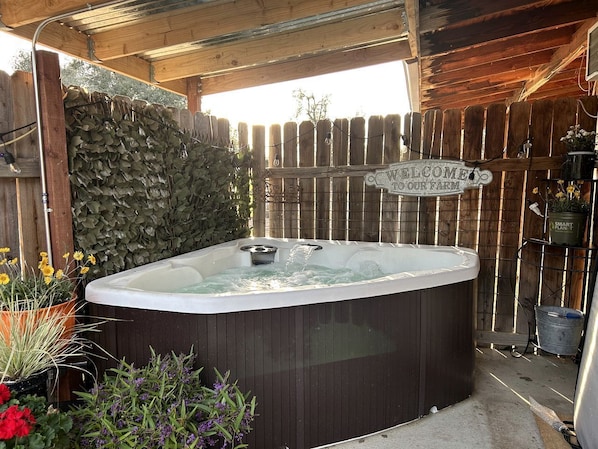A hot tub under a wooden pergola with a 'WELCOME TO OUR FARM' sign, surrounded by plants and a wooden fence.