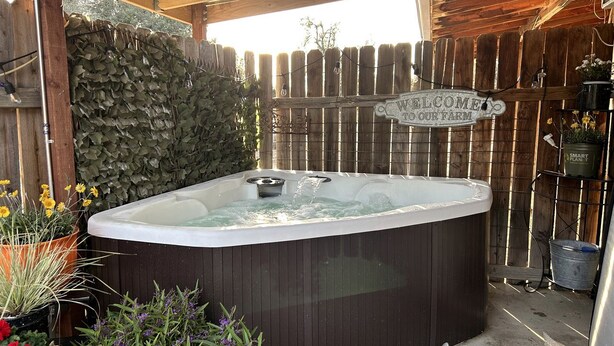 Un bain à remous sous une pergola en bois, agrémenté d’une pancarte indiquant « BIENVENUE À NOTRE FERME », entouré de plantes et d’une clôture en bois.