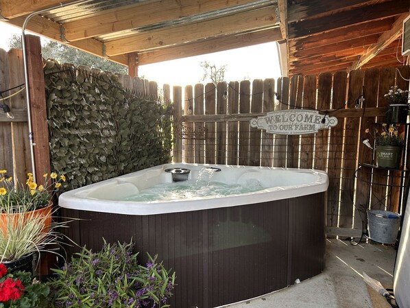 Un bain à remous sous une pergola en bois, agrémenté d’une pancarte indiquant « BIENVENUE À NOTRE FERME », entouré de plantes et d’une clôture en bois.