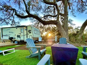 Een recreatiewagen met een kitchenette, twee Adirondack-stoelen, een picknicktafel en een grote boom op de achtergrond.