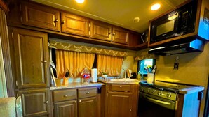A compact kitchen with wooden cabinets, a stove, a microwave, and a window with curtains.