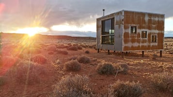 A small, single-story building with a glass door and windows, situated in a desert landscape with sparse vegetation.