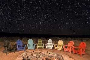 Eine Reihe Adirondack-Stühle sind um eine Steinkaminfeuerstelle unter einem sternenübersäten Nachthimmel angeordnet.
