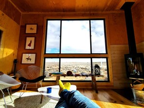 Una habitación con un ventanal grande, una guitarra, una silla, una mesa y un televisor.