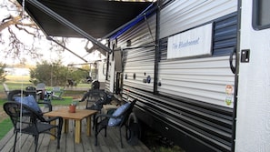 A covered outdoor area with a dining table and chairs, a sign reading 'The Bluebonnet,' and a trailer with a metal exterior. - Cozy Caravan in Texas for Romantic Getaways to Nature (Dripping Springs)