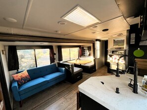 Interior de una caravana compacta, con un sofá azul, una pequeña mesa de comedor, una kitchenette y una ventana con vistas al exterior.