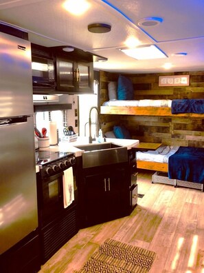 A compact kitchen with a stove, sink, and microwave, a bunk bed with pillows, and a wooden wall with a 'COZY' sign. - Cozy Caravan in Texas for Romantic Getaways to Nature (Dripping Springs)