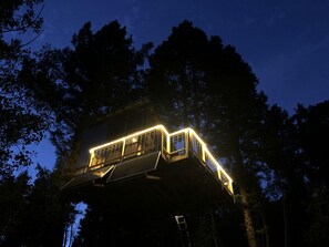 Une cabane perchée dans un arbre, dotée d’une terrasse en bois et de rampes, entourée d’arbres.