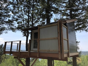 Ein hölzernes Baumhaus mit Balkon, das in einen Baum eingelassen ist und auf einen bewaldeten Bereich blickt.