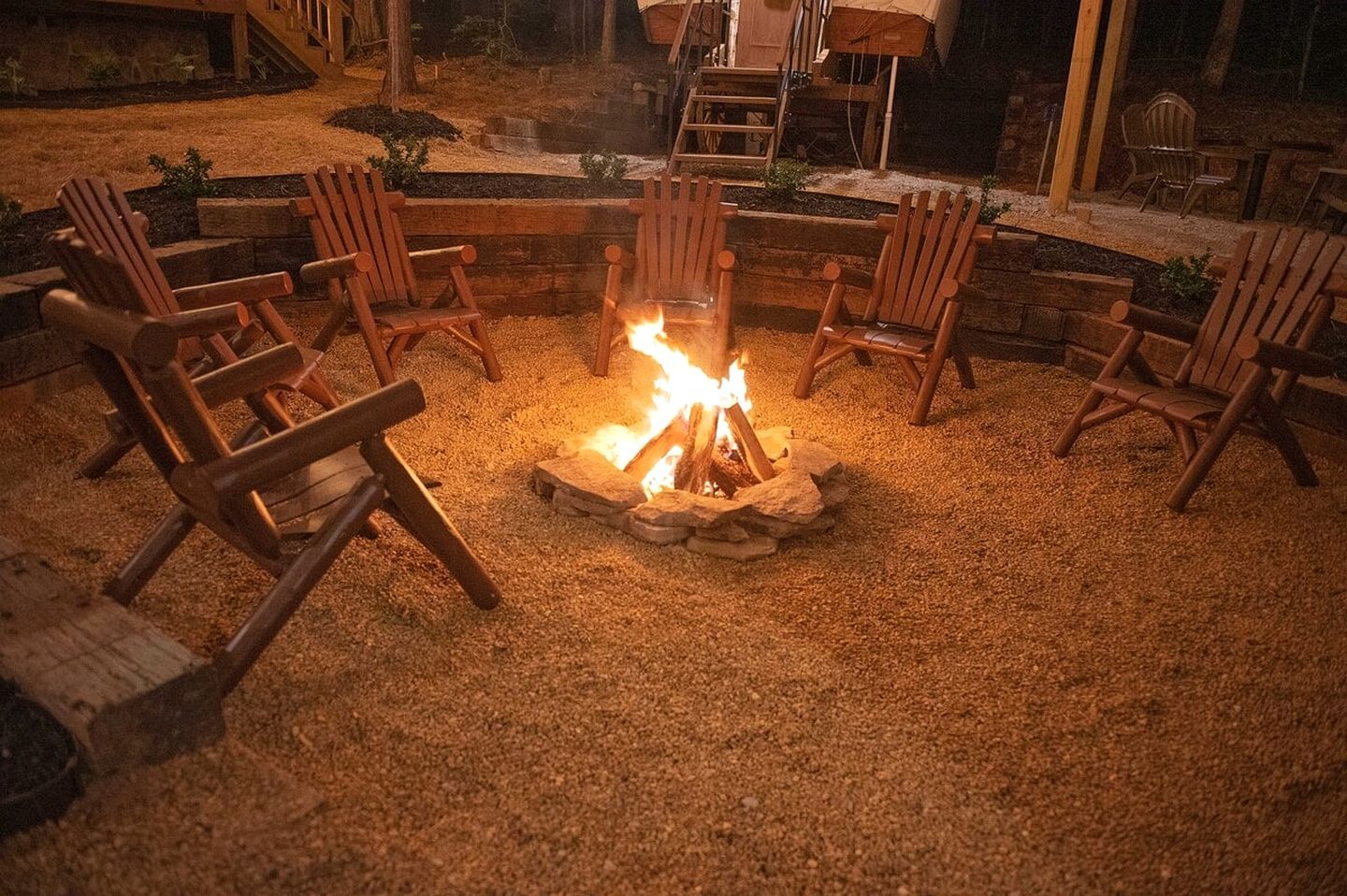 A fire pit with logs surrounded by wooden chairs in a gravel area.