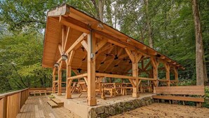 En träbeklädd paviljong med en grund av sten, omgiven av ett trädäck och bänkar, belägen i ett skogsområde.