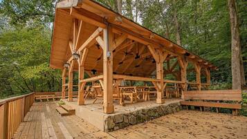 A wooden pavilion with a stone foundation, surrounded by a wooden deck and benches, set in a forested area.