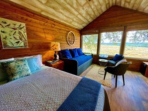 A wooden cabin bedroom with a bed, a sofa, a chair, and a large window. - Delightful Dripping Springs Rental for a Tranquil Getaway in Texas (Dripping Springs)