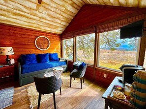 A cozy cabin interior with a wooden ceiling, a blue sofa, two chairs, a small table, and a view of the outdoors through large windows. - Delightful Dripping Springs Rental for a Tranquil Getaway in Texas (Dripping Springs)