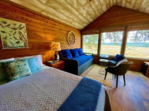 A wooden cabin bedroom with a bed, a sofa, a chair, and a large window.
