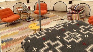 Una habitación con tienda de campaña, sillas naranjas, una pequeña mesa con libros, un tapete estampado y una cama con manta geométrica.