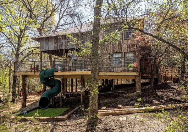 A rustic cabin with a wooden deck, a slide, and a fenced area.