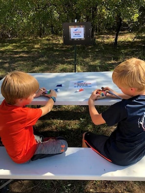 Twee kinderen schilderen aan een tafel buiten, met een bord in de achtergrond.