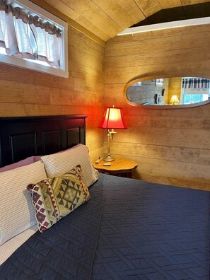 A bedroom with a bed, a nightstand with a lamp, a window with curtains, and a mirror on the wall.