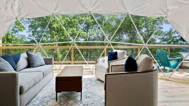 Un espacio al aire libre con carpas, un sofá, un reposapiés y sillas, una terraza de madera y vistas a árboles.