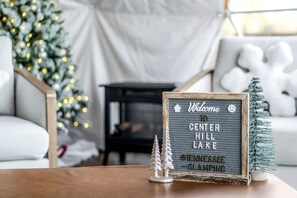 Un panneau d’accueil encadré avec l’inscription « Bienvenue au lac Center Hill, Tennessee – Expérience de camping de luxe », un petit sapin de Noël et un coin salon confortable avec un canapé blanc et une petite table.