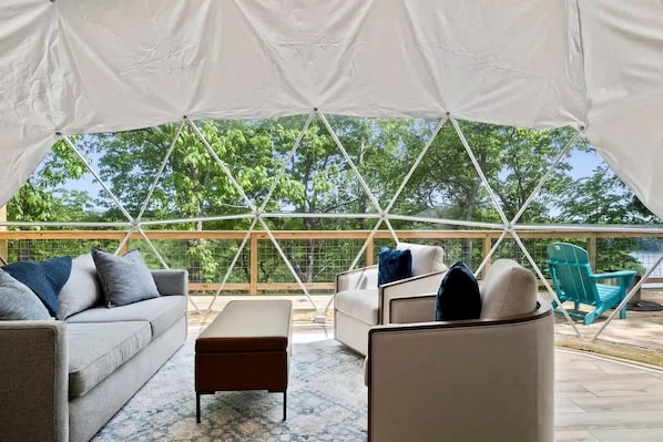 A tented outdoor area with a sofa, ottoman, and chairs, a wooden deck, and a view of trees. - Fully-Equipped Lake Dome with Beautiful Interior and Exterior in Tennessee (Smithville)