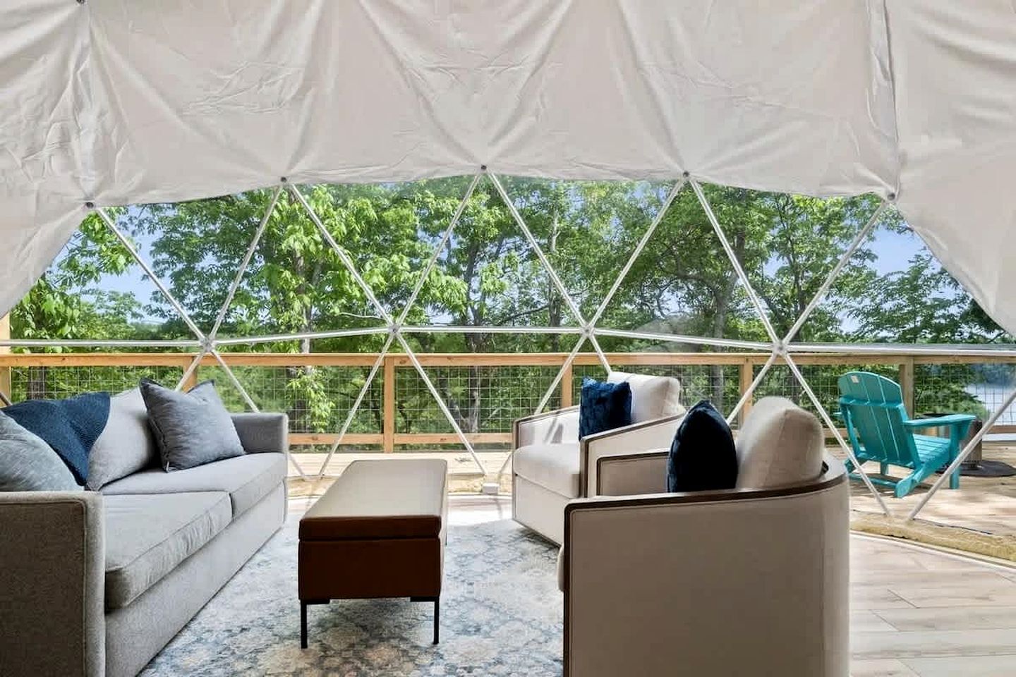 Ein überdachter Außenbereich mit einer Couch, Hocker und Sesseln, einer Holzterrasse und Blick auf Bäume.