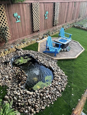 A backyard with a pond, wooden deck, and blue Adirondack chairs.