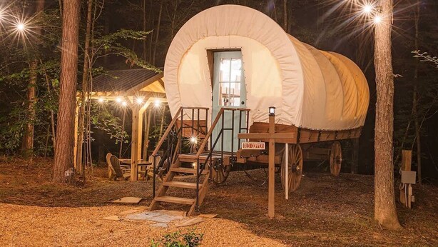 Uma tenda estilo carroça coberta com uma escadaria de madeira que leva a uma porta, rodeada por árvores e iluminação externa.