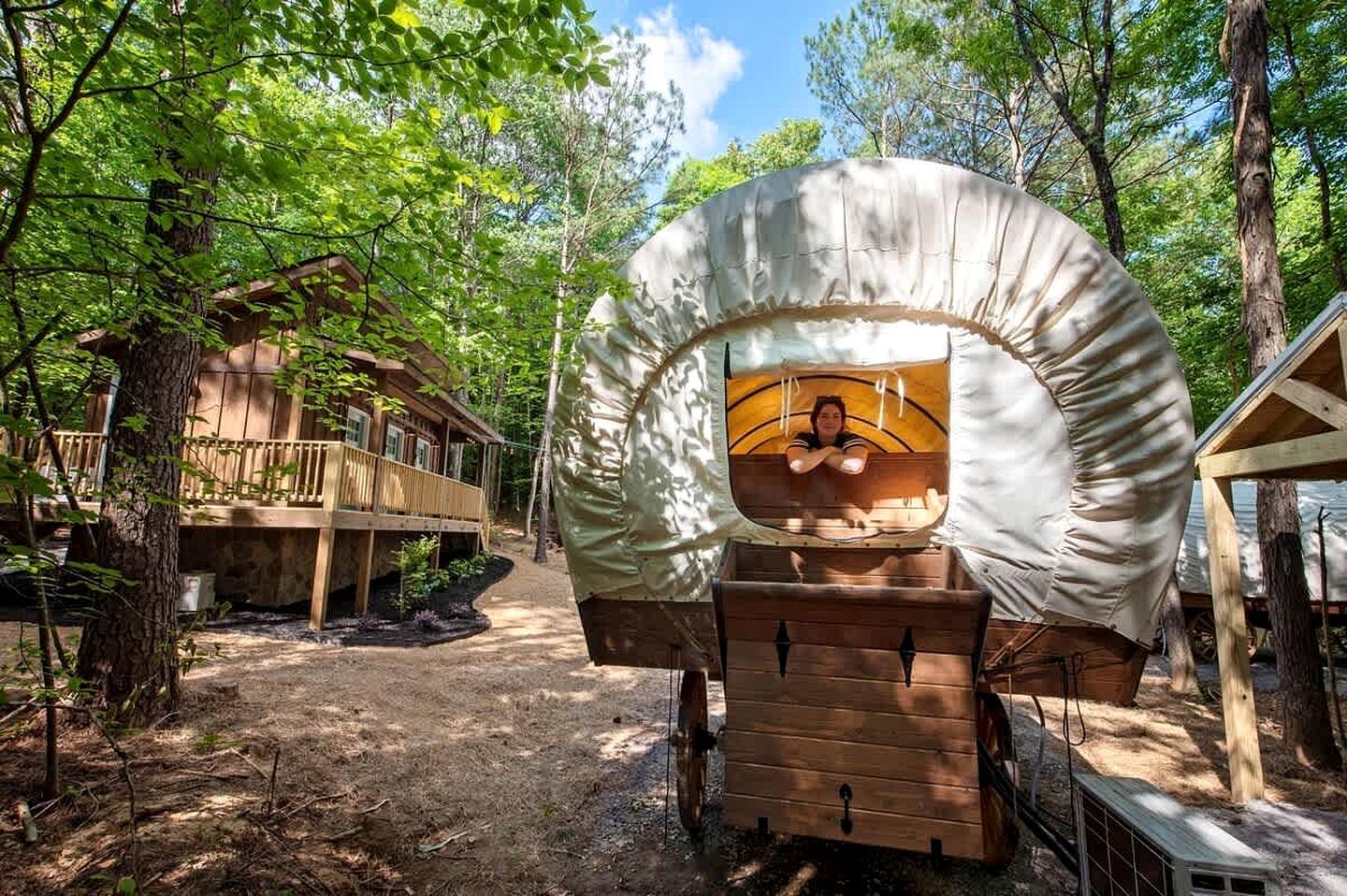 A covered wooden carriage with a person inside, surrounded by a forest and a wooden building with a balcony.