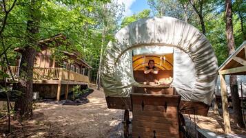 A covered wooden carriage with a person inside, surrounded by a forest and a wooden building with a balcony.