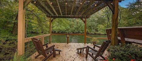 A wooden deck with two chairs and a bench, overlooking a body of water, surrounded by greenery.