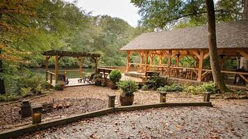 A covered outdoor area with picnic tables and a gazebo, surrounded by trees and a body of water.