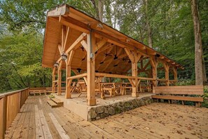 A wooden pavilion with a stone foundation, surrounded by a wooden deck and benches, set in a forested area.