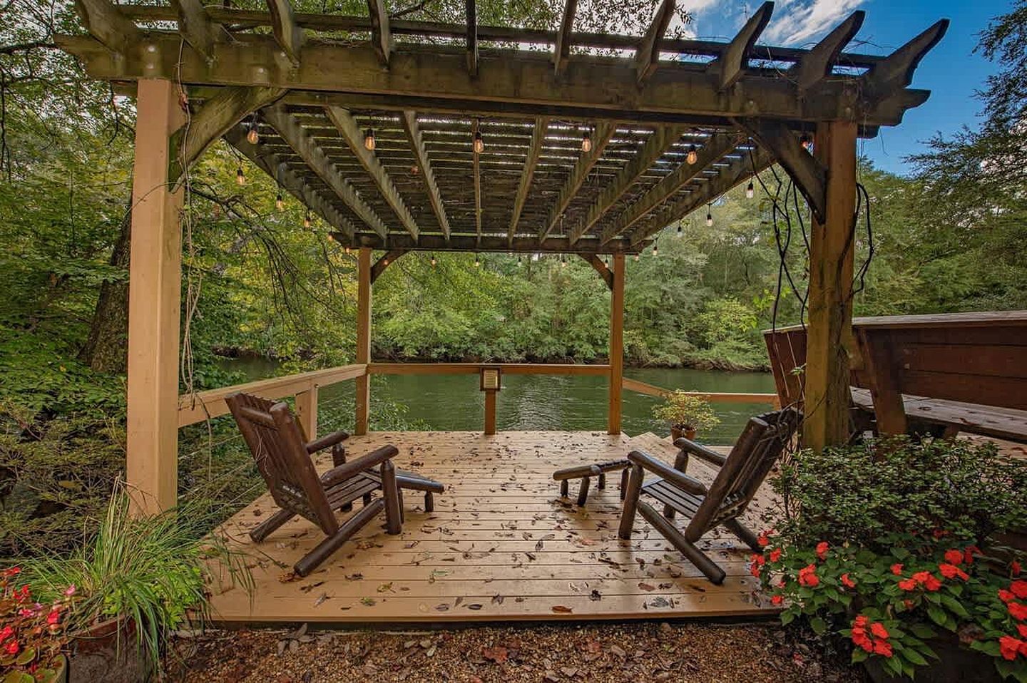 A wooden deck with two chairs and a bench, overlooking a body of water, surrounded by greenery.