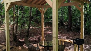 Un pabellón de madera con una mesa y sillas, rodeado de un bosque.