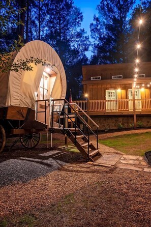 A rustic wooden cabin with a covered wagon attached, a staircase leading to the wagon, and a stone wall with windows.
