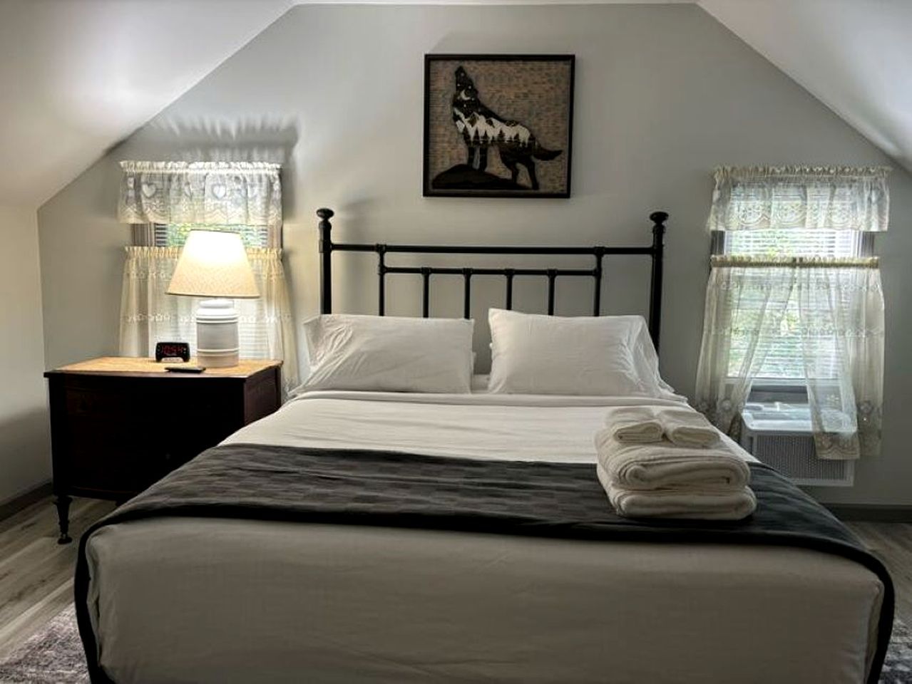 A bedroom with a bed, a nightstand, a lamp, a framed picture, and two windows with curtains.