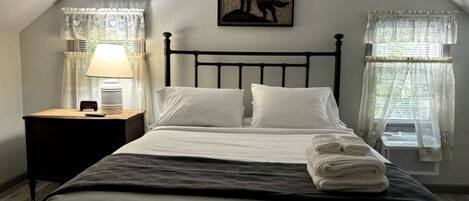 A bedroom with a bed, a nightstand, a lamp, a framed picture, and two windows with curtains.