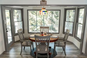 A dining area with a round table, four chairs, a stained glass chandelier, and large windows with a view of trees.