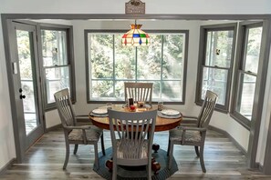 A dining area with a round table, four chairs, a stained glass chandelier, and large windows with a view of trees.