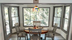 A dining area with a round table, four chairs, a stained glass chandelier, and large windows with a view of trees.