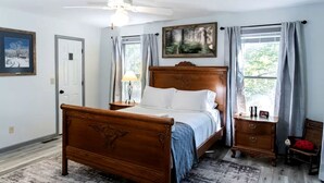 A bedroom with a wooden bed, a nightstand, a lamp, a ceiling fan, a painting, and a window with curtains.