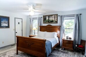 A bedroom with a wooden bed, a nightstand, a lamp, a ceiling fan, a painting, and a window with curtains.