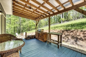 A covered outdoor deck with a table and chairs, a wooden cabinet, and a view of a forested hillside.